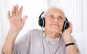An elderly woman with grey hair is wearing a light sweater, and a pearl necklace. she has one hand touching the headphones she is wearing and one hand in the air, as she appears to be enjoying what she is hearing.