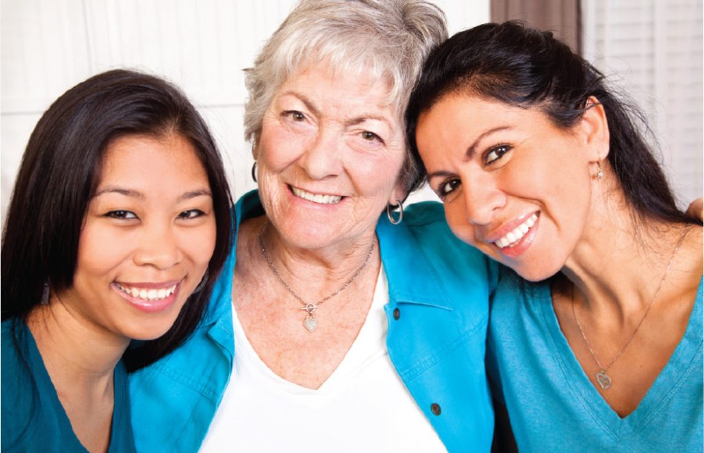 A smiling woman with grey hair has her arms around two women with long dark hair on either side of her. Everyone is smiling into the camera.