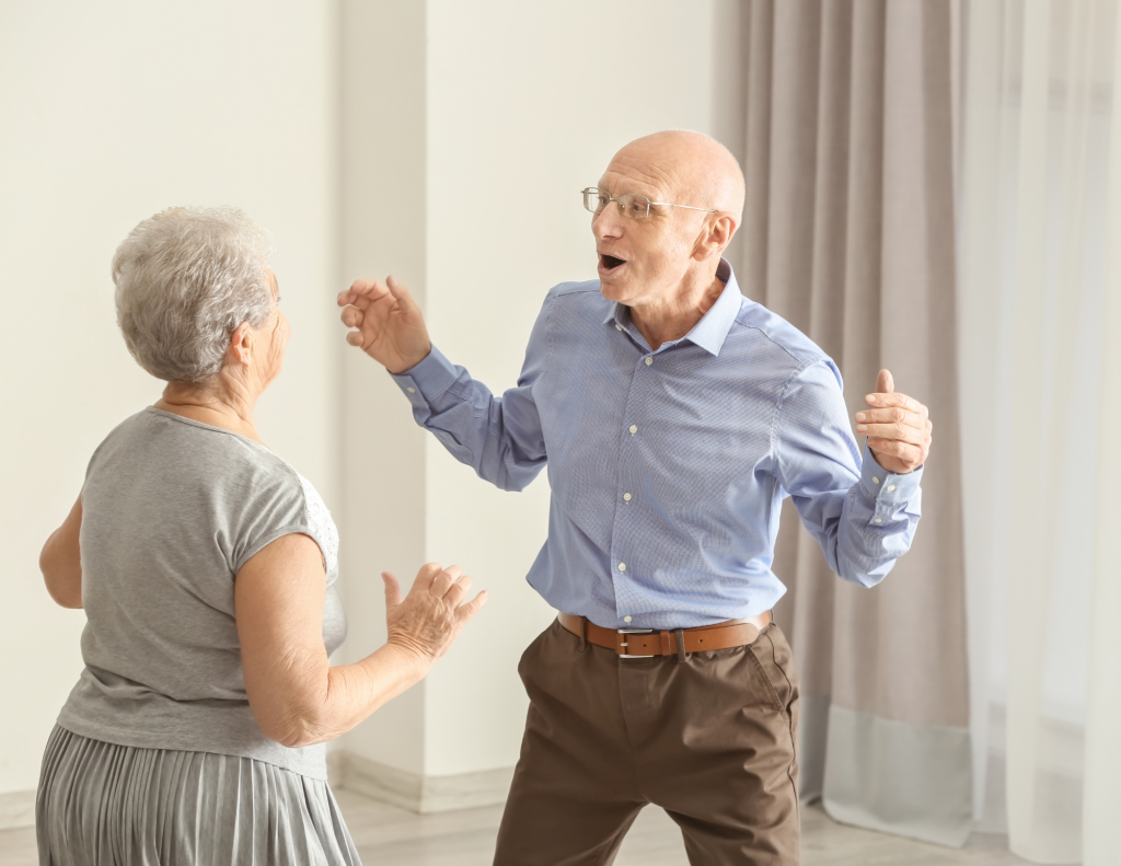 An older, think, bald man with glasses and a blue collared shirt and brown pants with a brown leather belt  and a woman with grey hair, a grey blouse and a grey skirt and facing each other and dancing with their arms moving but not touching each other.