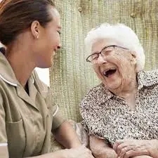 A young woman with dark hair pulled back and wearing a tan blouse is laughing with an elderly woman with short grey hear, glasses, and a brown and tan patterned blouse.