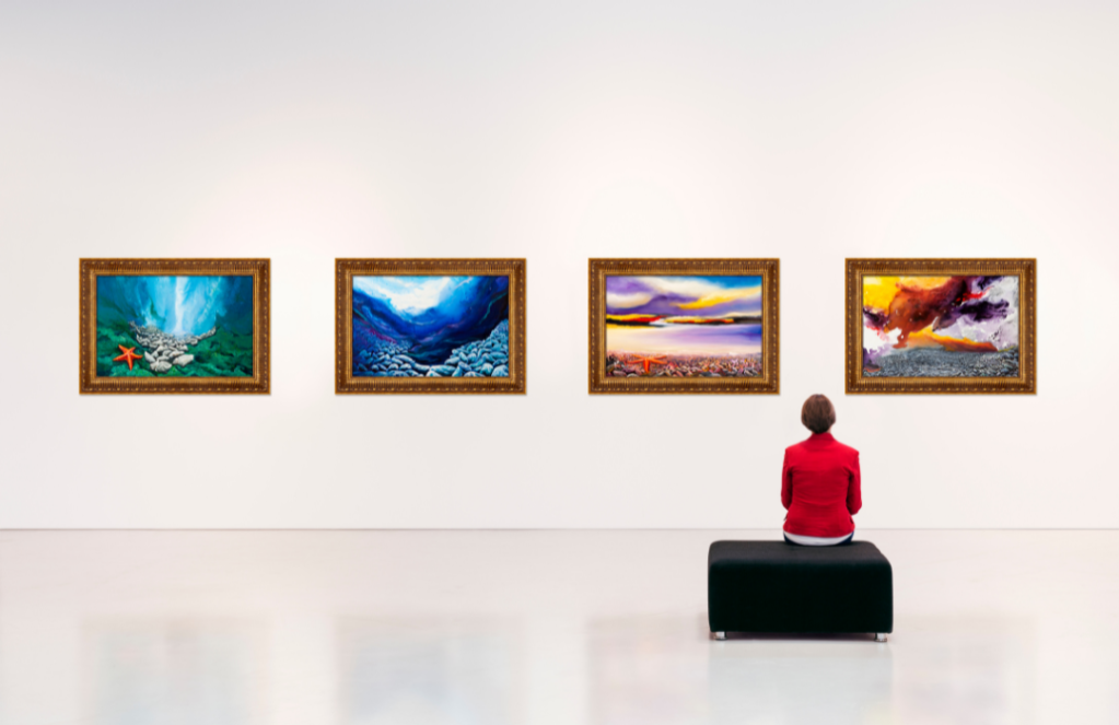 A with short brown hear and wearing a red shirt is sitting on a black stool facing a white wall with various framed pieces of art hanging horizontally on it.